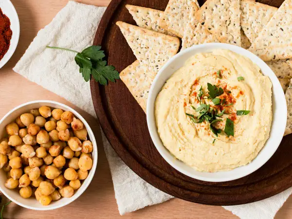 Un tazón de hummus cremoso, decorado con perejil y pimentón, junto a garbanzos y galletas saladas. ¡Perfecto para tu hummus receta!