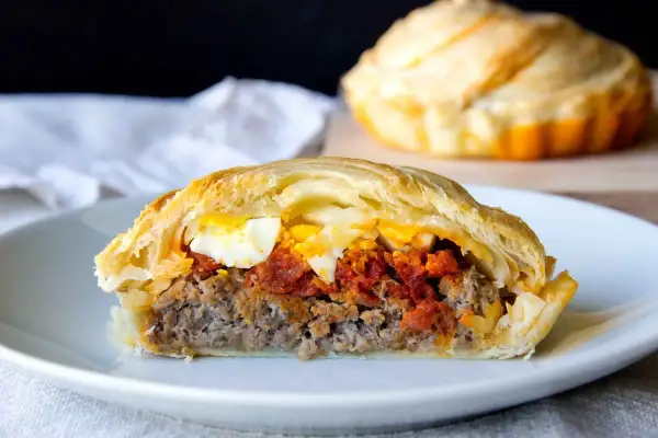 Un trozo de pastel de carne murciano, relleno de carne picada, tomate y huevo duro, envuelto en hojaldre dorado.