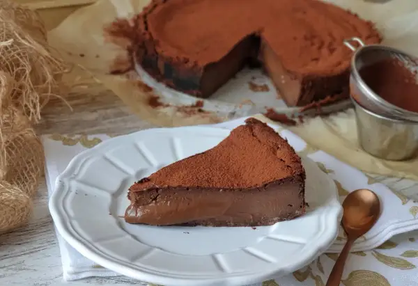 Una porción de tarta de chocolate, cubierta de cacao en polvo, reposa en un plato blanco. El resto de la tarta, también espolvoreada, se ve al fondo.