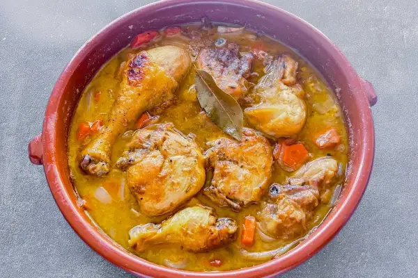Sabroso pollo en salsa con trozos dorados, zanahorias y laurel en una cazuela de barro.