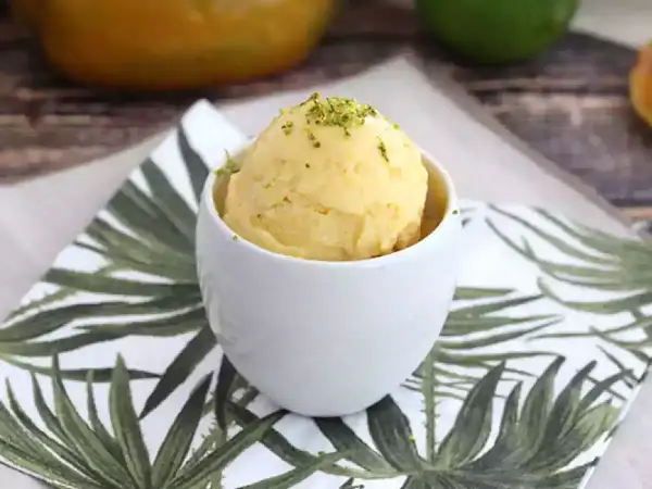 Una bola de sorbete de mango, refrescante y deliciosa, servida en una taza blanca sobre una servilleta con hojas tropicales.