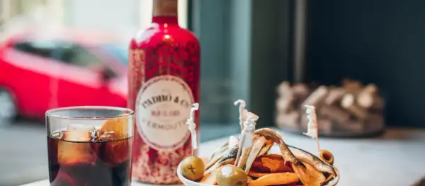 Un vaso de vermut con hielo y naranja, junto a una botella roja de Padró & Co. Vermouth y un plato de patatas y anchoas. ¡Perfecto para clases de vermut!