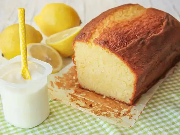 Bizcocho de yogur casero, dorado y esponjoso, acompañado de limones frescos y un vasito de yogur.