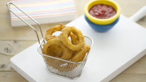Crujientes aros de cebolla dorados en una cesta metálica, listos para ser sumergidos en kétchup.