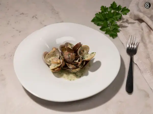 Un plato blanco con almejas en salsa verde, adornado con perejil fresco. Un tenedor y una servilleta completan la escena.