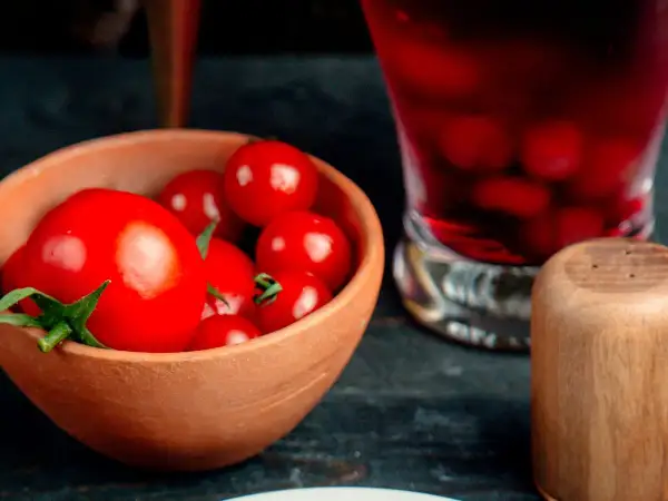 Un cuenco de barro lleno de tomates rojos brillantes, listos para un delicioso tartar de tomate. A su lado, un vaso con bebida roja y un salero de madera.